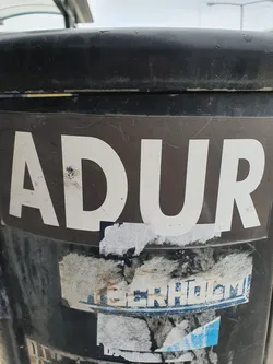 A sticker with the word ADUR in white capital letters on a dark brown background. The sticker is affixed to a dark-colored cylindrical object, possibly a trash can.  There are other damaged stickers underneath it.