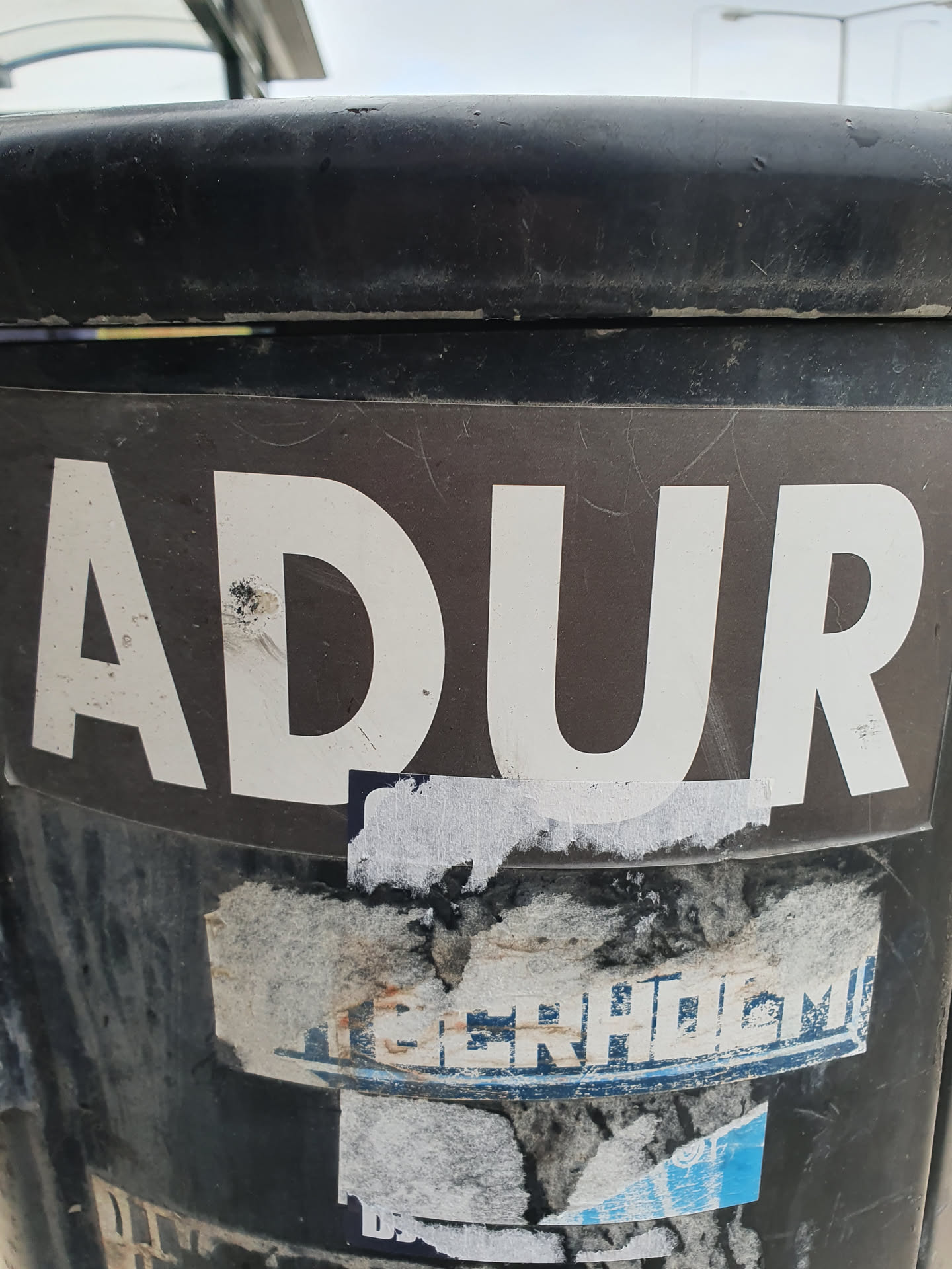 A sticker with the word ADUR in white capital letters on a dark brown background. The sticker is affixed to a dark-colored cylindrical object, possibly a trash can.  There are other damaged stickers underneath it.