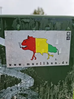 A sticker featuring a cartoonish dog-like creature smoking a cigarette. The creature is colored in red, yellow, and green, resembling a flag. The background is white and wet, with water droplets visible.  A website address and social media icons (Facebook and Instagram) are present on the sticker.