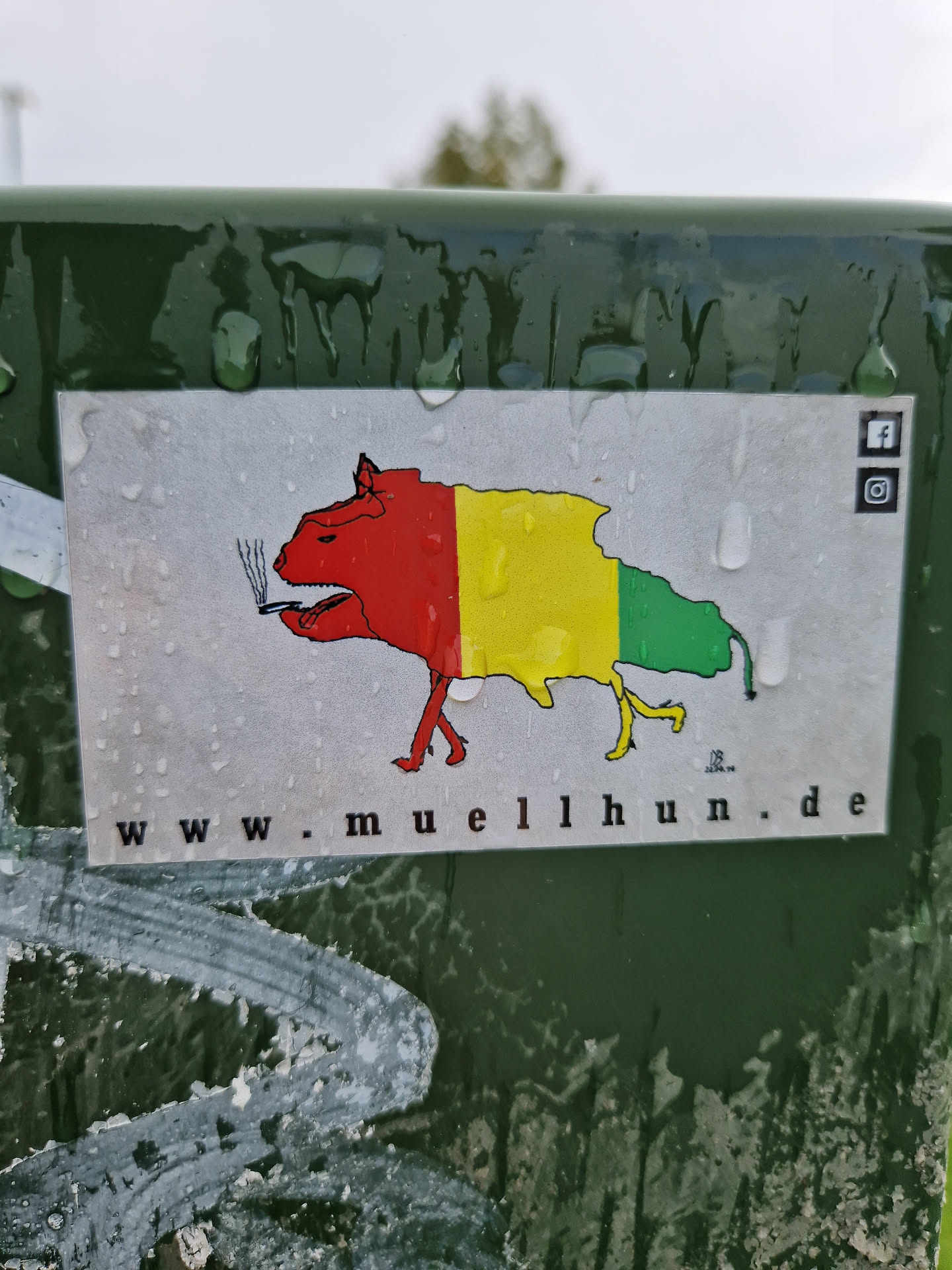 A sticker featuring a cartoonish dog-like creature smoking a cigarette. The creature is colored in red, yellow, and green, resembling a flag. The background is white and wet, with water droplets visible.  A website address and social media icons (Facebook and Instagram) are present on the sticker.