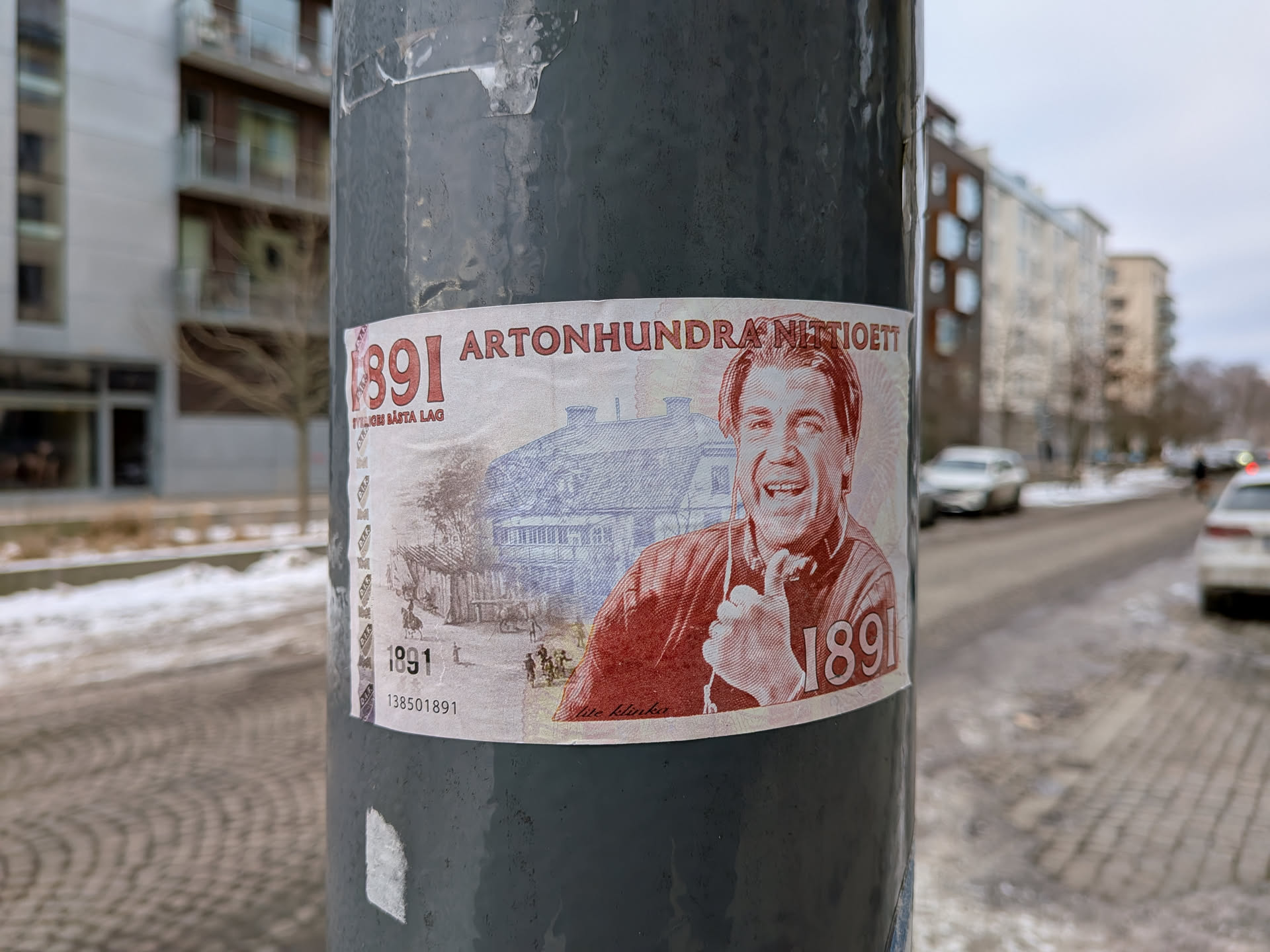 The sticker is designed to resemble an old banknote, predominantly in off-white, red-orange, and blue-grey tones. It features a prominent portrait of a smiling man, possibly a caricature, rendered in a red-orange, engraved style. He wears a collared shirt with the number '1891' on it and appears to be holding something near his chest. Behind him, in a blue-grey, finely detailed engraving style, is an illustration of a large, traditional building, possibly a farm or manor, with smaller figures and horses in the foreground. Large red numbers '1891' are visible on the left side, with the text 'SVERIGES BÄSTA LAG' beneath them. At the top, 'ARTONHUNDRA NITTIOETT' is written in red. Smaller '1891' and a numerical sequence '138501891' are also present at the bottom left. The sticker has a slightly worn appearance, adhering to a dark grey pole.