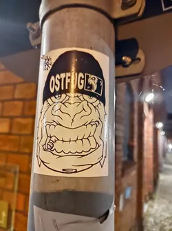 A monochrome sticker featuring a cartoonish, angry-looking creature wearing a beanie with the word "OSTFUG" written on it. The creature appears to be smoking a cigar. The art style is simple yet effective, with bold lines and stark contrasts.