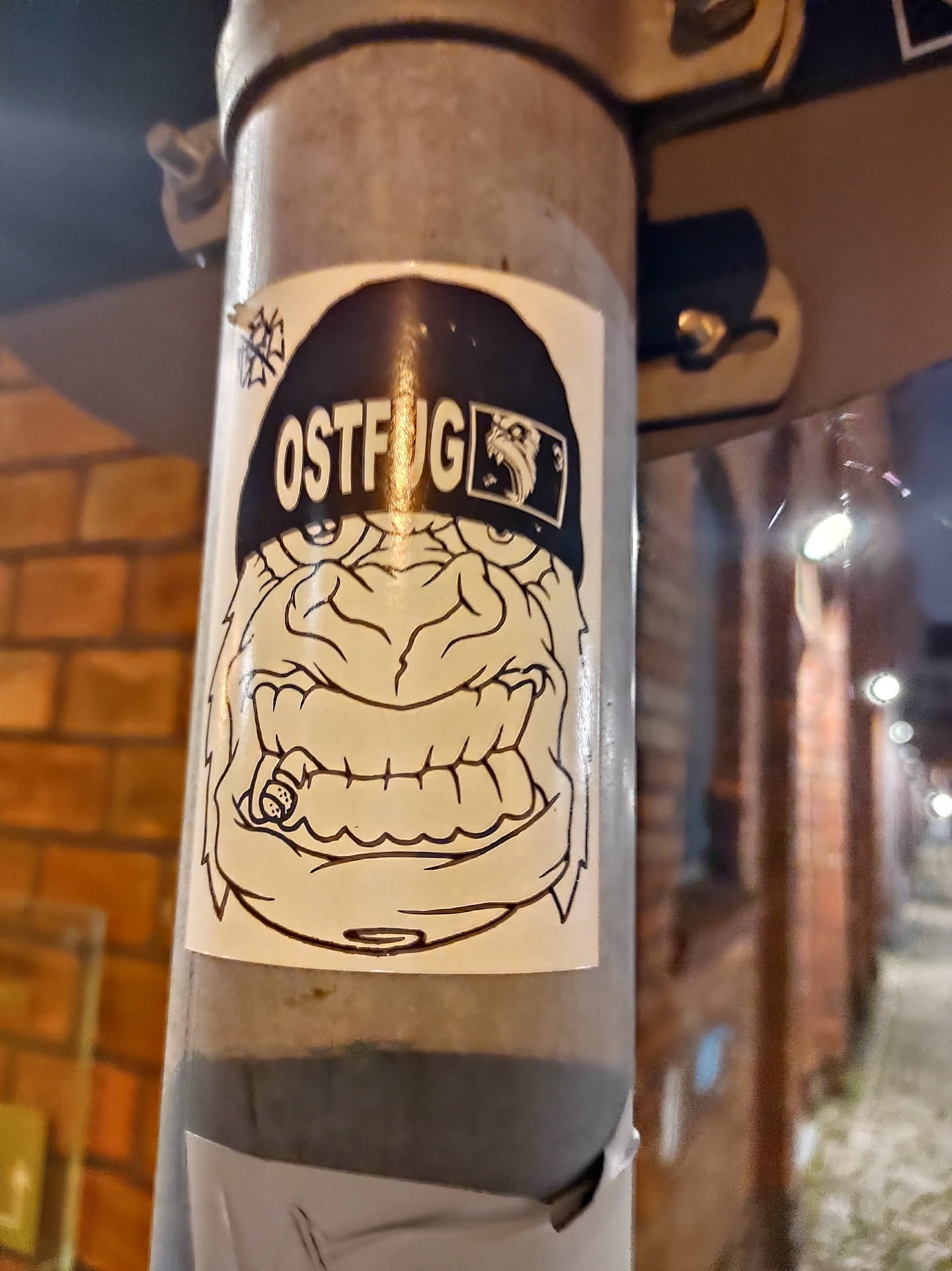 A monochrome sticker featuring a cartoonish, angry-looking creature wearing a beanie with the word "OSTFUG" written on it. The creature appears to be smoking a cigar. The art style is simple yet effective, with bold lines and stark contrasts.