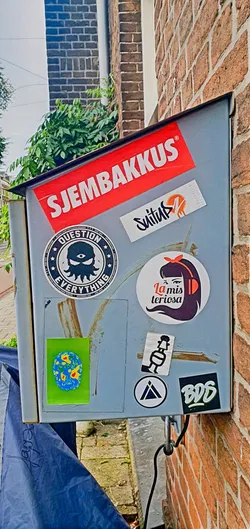 The image features a grey metal box fixed to a brick wall, covered with various stickers. Prominently displayed is a red sticker with white text "SJEMBAKKUS". Below, a round sticker with an eye and tentacle design reads "QUESTION EVERYTHING". Other stickers include a logo resembling a paint spray, a sticker with the text "La mis teriosa" alongside a graphic of a woman with headphones, and a small square sticker featuring a schematic drawing. The overall impression is a collage of diverse graphics and texts on a weathered metal surface.