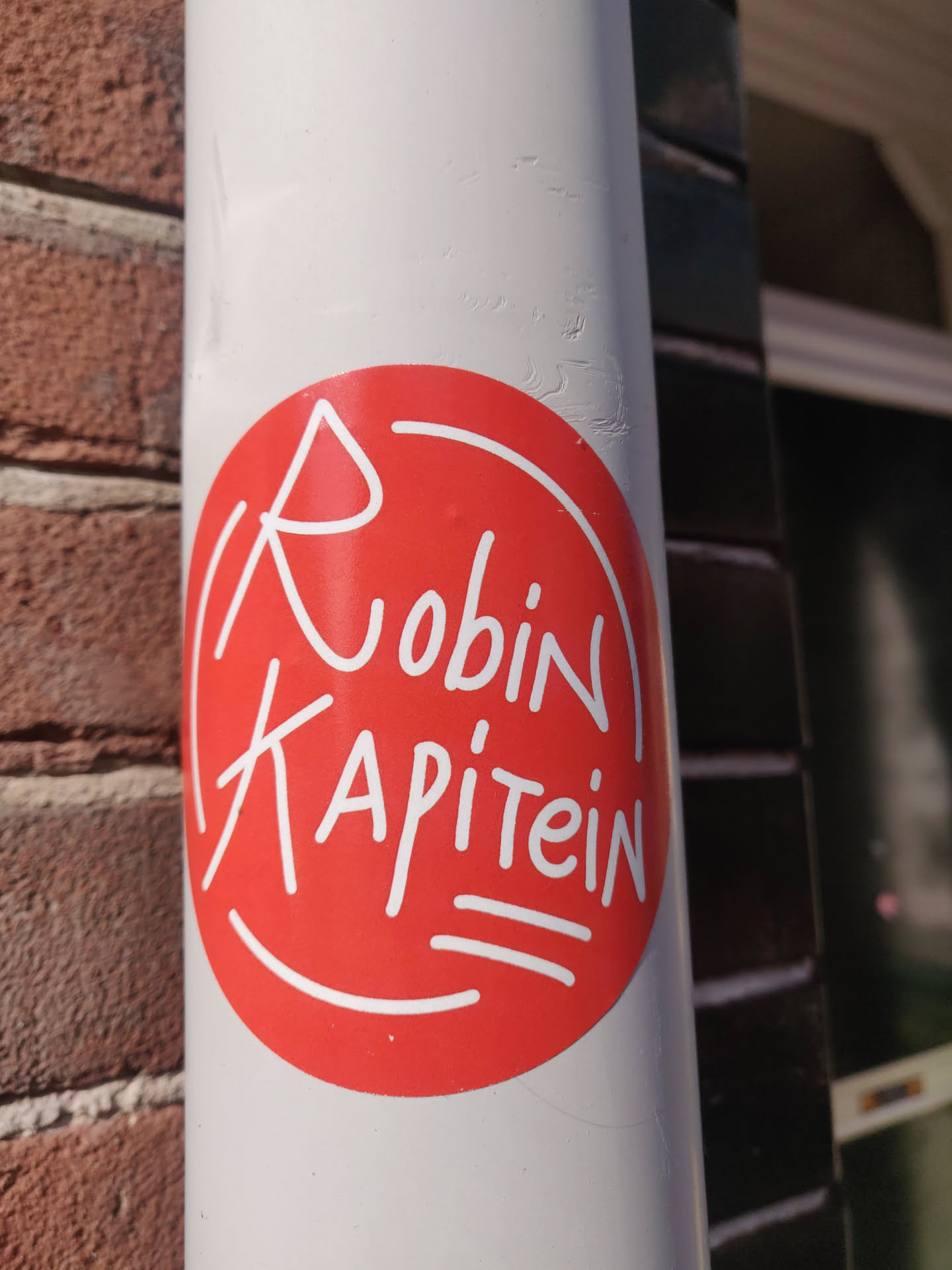 A red circular sticker with the words 'Robin Kapitein' written in a stylized white script. The design is simple yet eye-catching. The sticker is affixed to a white cylindrical surface, possibly a pipe or drainpipe.