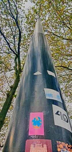 The image features a tall, cone-shaped structure with several stickers on its surface. The focused sticker is a pink rectangle with a blue and orange abstract design resembling a pixelated skull or flower. The structure is surrounded by trees with autumn leaves.