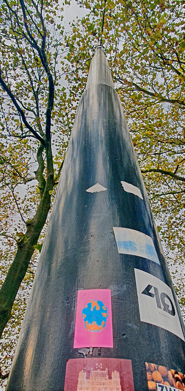 The image features a tall, cone-shaped structure with several stickers on its surface. The focused sticker is a pink rectangle with a blue and orange abstract design resembling a pixelated skull or flower. The structure is surrounded by trees with autumn leaves.