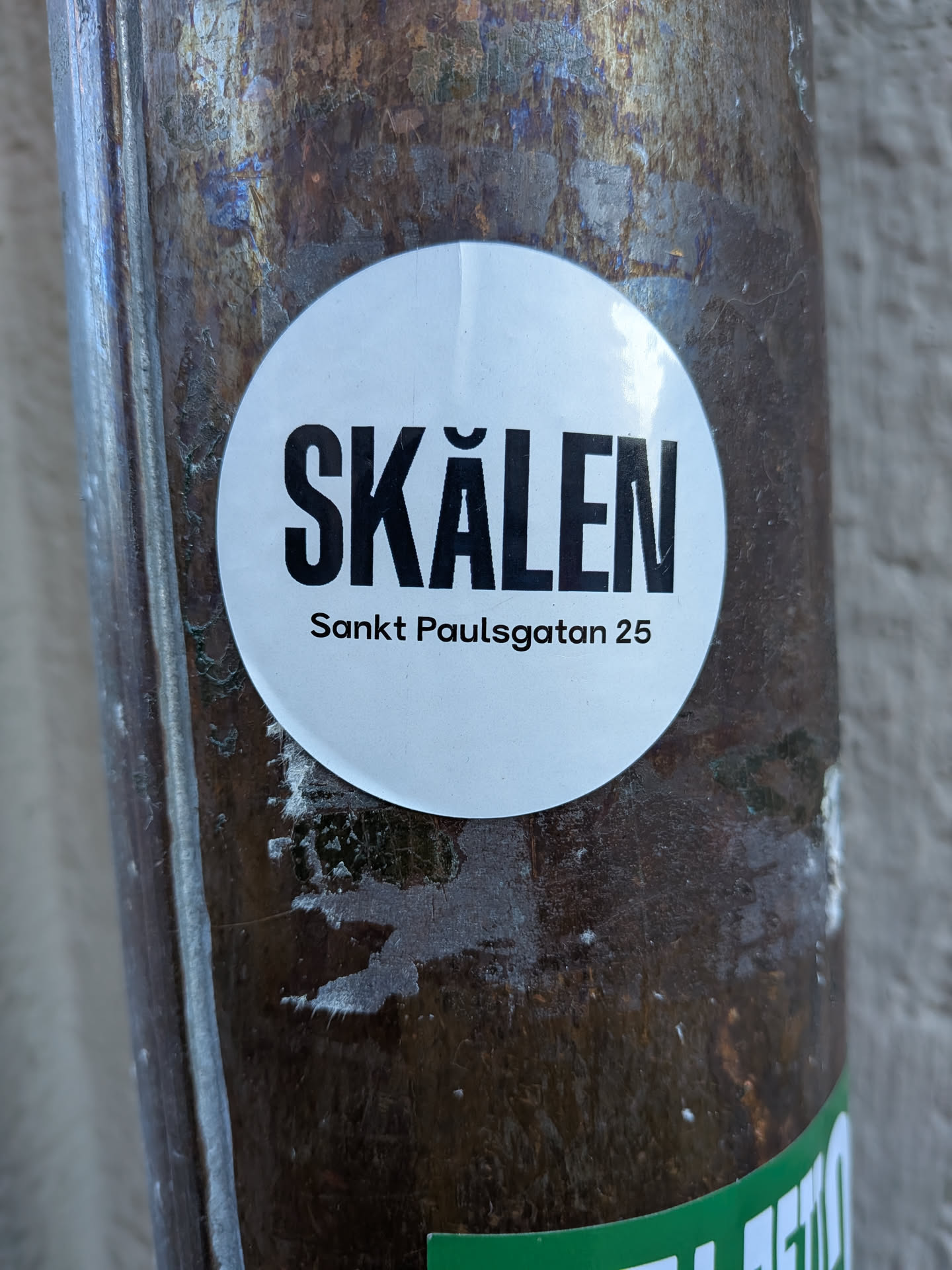A circular white sticker with bold black text, adhered to a textured, dark, possibly metallic or rusty surface. The main text 'SKÅLEN' is prominently displayed in large, uppercase, sans-serif font. Below it, in a smaller, regular sans-serif font, is the address 'Sankt Paulsgatan 25'. The sticker shows slight wear and minor creasing around its edges, indicating it's a physical sticker on an outdoor surface.