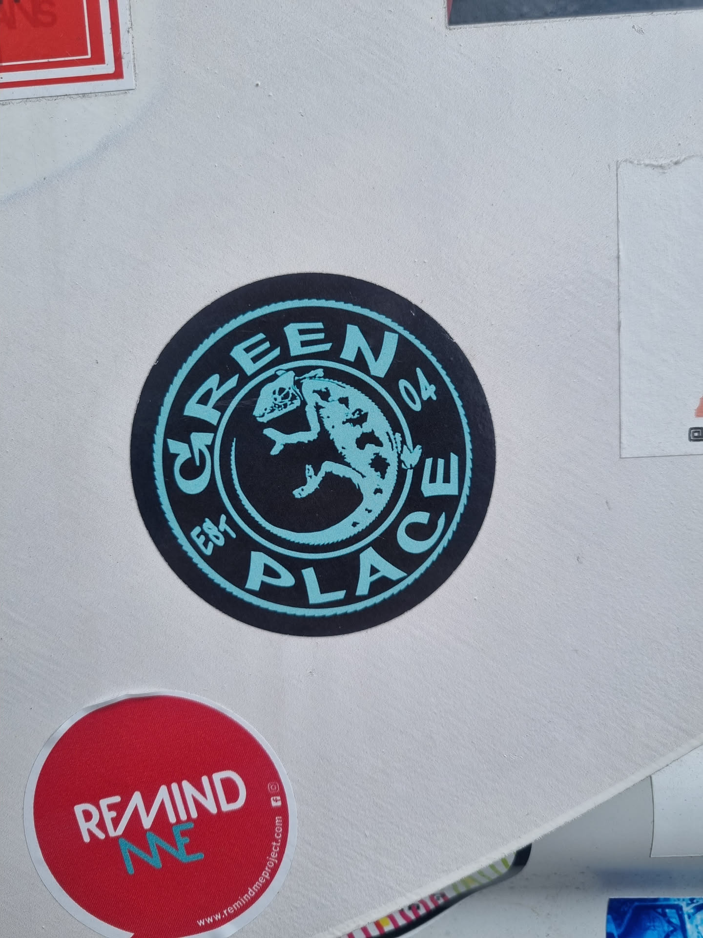 Round black and teal sticker with the words \"Green Place 04\" written around a central illustration of a gecko or lizard. The style is simple and slightly vintage.