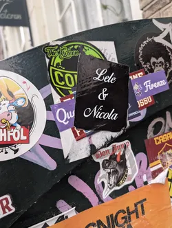 A rectangular black sticker with white cursive text "Lele & Nicola" is adhered to a surface covered in various other stickers. The surrounding stickers have different designs and colors, creating a cluttered and textured background.