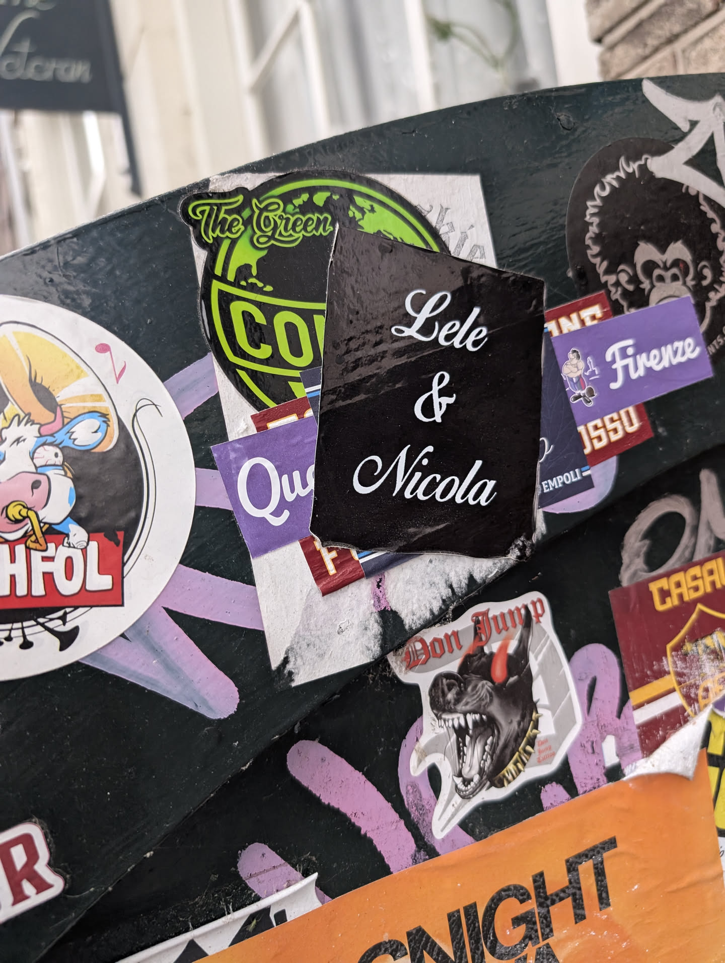 A rectangular black sticker with white cursive text "Lele & Nicola" is adhered to a surface covered in various other stickers. The surrounding stickers have different designs and colors, creating a cluttered and textured background.