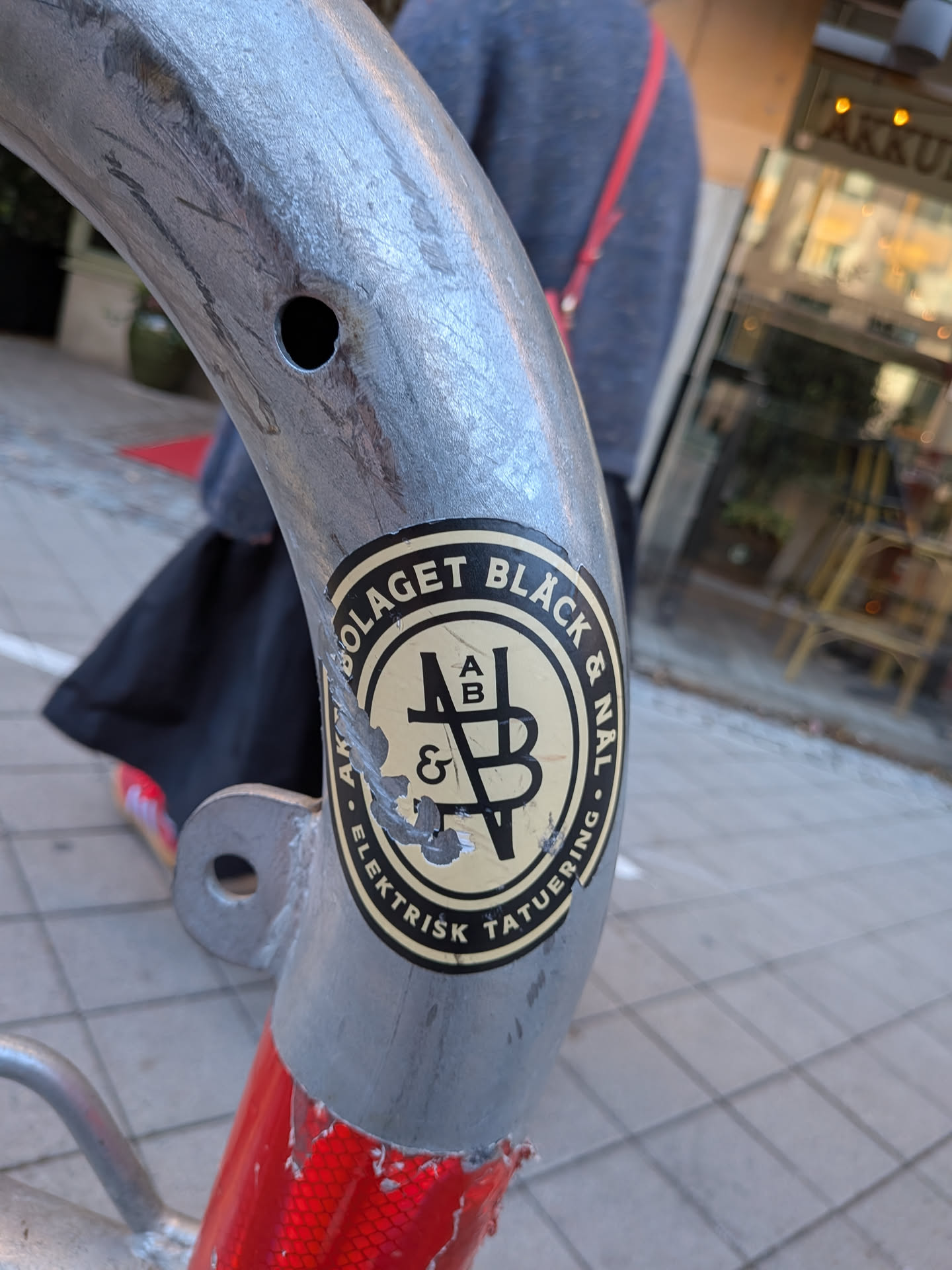 An oval-shaped sticker with a cream-white background and black design, adhered to a curved, galvanised metal post. The outer border features bold circular text in black reading 'BOLAGET BLÄCK & NÅL' across the top and 'ELEKTRISK TATUERING' along the bottom. Inside an inner oval border is a stylized monogram featuring interconnected geometric letters and symbols, including 'A', 'B', and '&'. The left edge of the sticker shows some minor wear and scratching.
