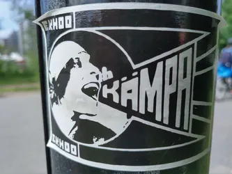 A black and white sticker featuring a stylized illustration of a person shouting into a megaphone with the word "KÄMPA" (Swedish for "fight" or "struggle") prominently displayed.  The style is bold and graphic. The word "EXHOO" appears in the corners, possibly a brand or artist signature. The sticker is affixed to a dark-colored pole.