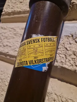A yellow sticker on a brown pipe. The sticker contains text in Swedish and some diagrams.  The text translates roughly to "Save Swedish Football! Scrap the conditional ladder." The years 2013, 2021, and 2027 are also visible, possibly indicating timeframes or goals.