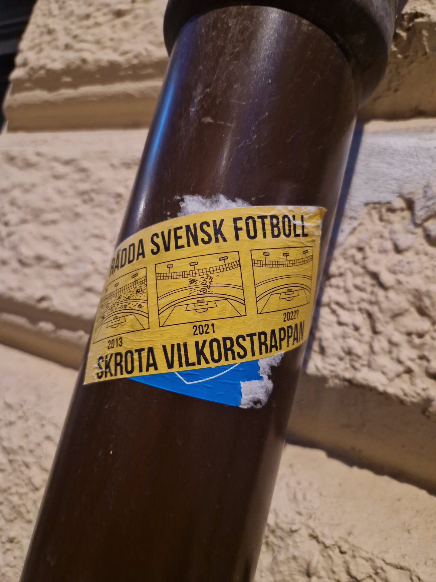 A yellow sticker on a brown pipe. The sticker contains text in Swedish and some diagrams.  The text translates roughly to "Save Swedish Football! Scrap the conditional ladder." The years 2013, 2021, and 2027 are also visible, possibly indicating timeframes or goals.