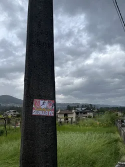 A rectangular sticker with a prominent red border, affixed to a dark, textured concrete pole. The design features a collage of elements associated with Sevilla FC. On the left, a gold laurel wreath encircles a crest with the letters 'SFC' in white on a red background. In the center, a black silhouette of a figure, possibly a bullfighter or a traditional dancer, is depicted. To the right, another shield-like crest with vertical red and white stripes is visible, topped by smaller text including 'LLAIMOMANO' and 'VIDA'. The bottom section of the sticker has a red background with the white text 'adictos al SEVILLA FC' in a bold, sans-serif font. The overall color scheme is red, white, black, and gold.