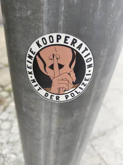A round sticker with a illustration of a woman putting a finger to her lips. The text on the sticker says: "Keine Kooperation mit der Polizei" which translates to: "No cooperation with the police"