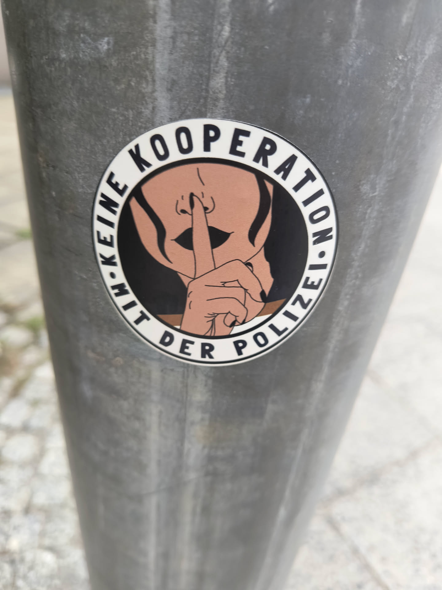 A round sticker with a illustration of a woman putting a finger to her lips. The text on the sticker says: "Keine Kooperation mit der Polizei" which translates to: "No cooperation with the police"