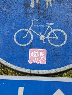 A pink square sticker with the text \"ACTIVE BOYZ\" in a stylized font is affixed to a weathered blue sign featuring a bicycle symbol.  The sign shows signs of age and wear. 