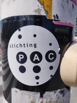 A black and white circular sticker with the word 'stichting' and the letters 'PAC' in the center, surrounded by various sized dots. The sticker is adhered to a pole that is covered in other layers of stickers and grime.