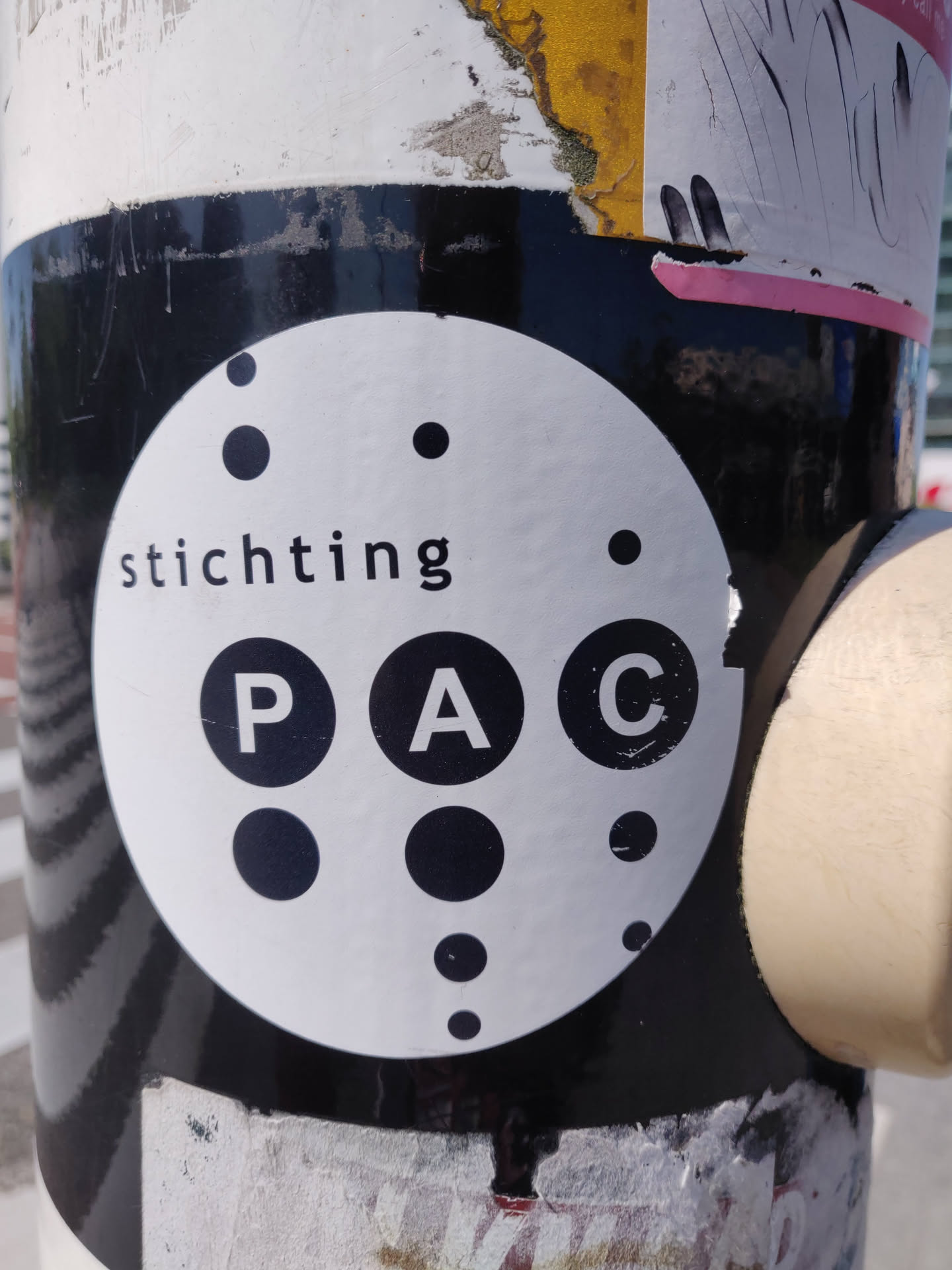 A black and white circular sticker with the word 'stichting' and the letters 'PAC' in the center, surrounded by various sized dots. The sticker is adhered to a pole that is covered in other layers of stickers and grime.