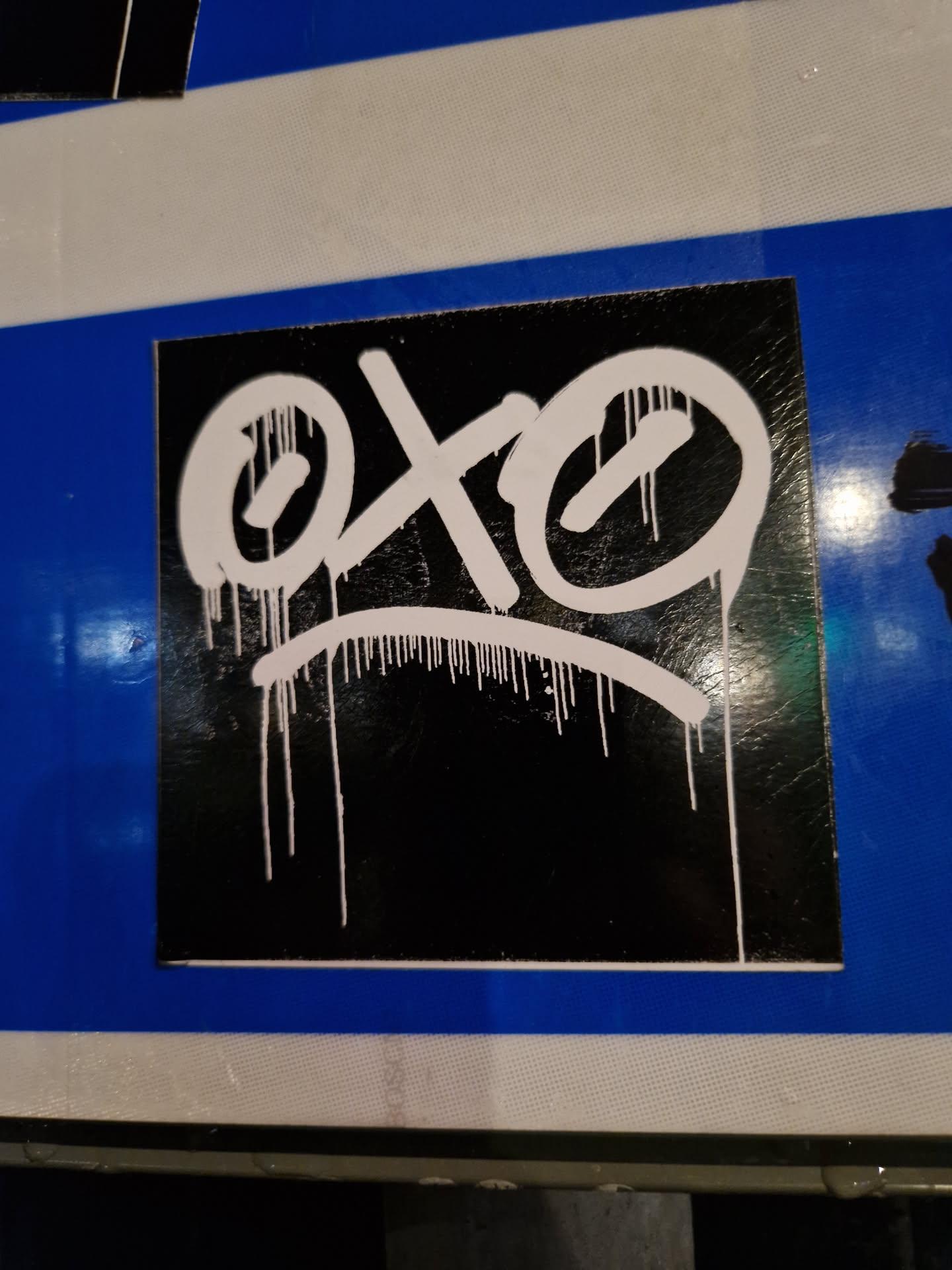 This square sticker features a graphic design in white paint on a black background. The design resembles a face with two large circular eyes and a simple curved line representing the mouth. White paint drips down from the figure, adding a dynamic feel. The sticker has a clean, glossy finish, and the graphic is bold and expressive, creating a striking visual contrast.