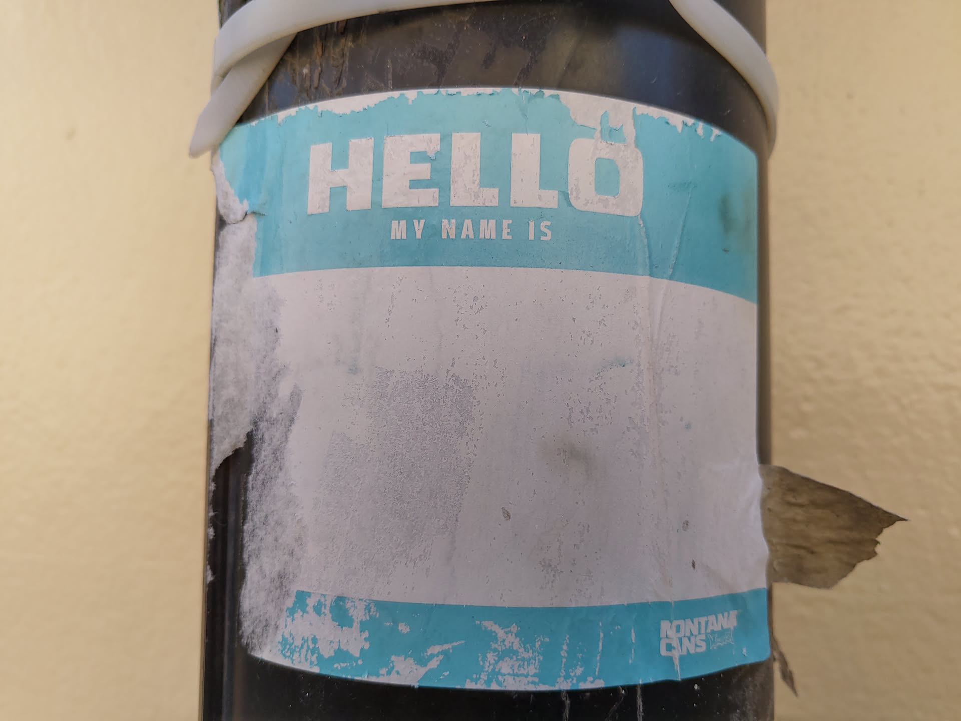 A light teal and white name tag sticker is affixed to a dark pole.  The sticker is peeling and worn, with significant damage and dirt. The top portion says "HELLO MY NAME IS" in a sans-serif font. The bottom portion is mostly blank but has a small amount of text visible at the bottom right, possibly a brand or logo.