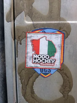 The sticker features a central square design with a distressed, light grey background. Within this square is an outline of a landmass, possibly a stylized map, filled with three vertical stripes: red on the left, white in the middle, and green on the right, reminiscent of the Italian flag. Below this graphic, in prominent white letters with a red outline and shadow, is 'MODO HOCKEY'. Beneath that, in smaller, red cursive-style text, is 'Örnsköldsvik'. The square is framed by thin green and red borders. This central element is placed on a larger, shield-shaped background, which is light blue and bordered by thin lines of red, yellow, and black. Partially visible at the bottom of the shield are the dark blue letters 'UCS'. The sticker is adhered to a metallic surface, which is stained with dark brown, muddy drips.