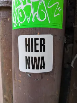A rectangular white sticker with slightly rounded corners, featuring two lines of bold, black, sans-serif capital letters. The top line reads 'HIER' and the bottom line reads 'NWA'. The sticker is applied to a rusty, dark brown metal pole, showing minor signs of wear and tear around its edges.