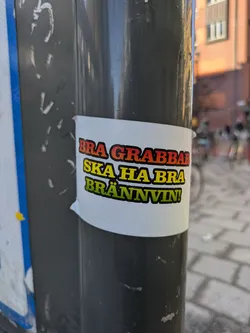 A sticker on a pole with Swedish text that reads "BRA GRABBAR SKA HA BRA BRÄNNVIN!", which translates to "GOOD GUYS SHOULD HAVE GOOD BOOZE!"