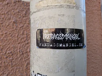 Street sticker A rectangular glossy black sticker adhered to a weathered, off-white painted metal pipe. At the center is a spiky, black-metal style logo in white that reads 'KLUBB FREDAGSMANGEL'. Beneath the logo, the website 'FREDAGSMANGEL.SE' is printed in a distressed white typeface.
