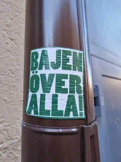 A green sticker with white text on a brown pole. The text says "BAJEN ÖVER ALLA!"