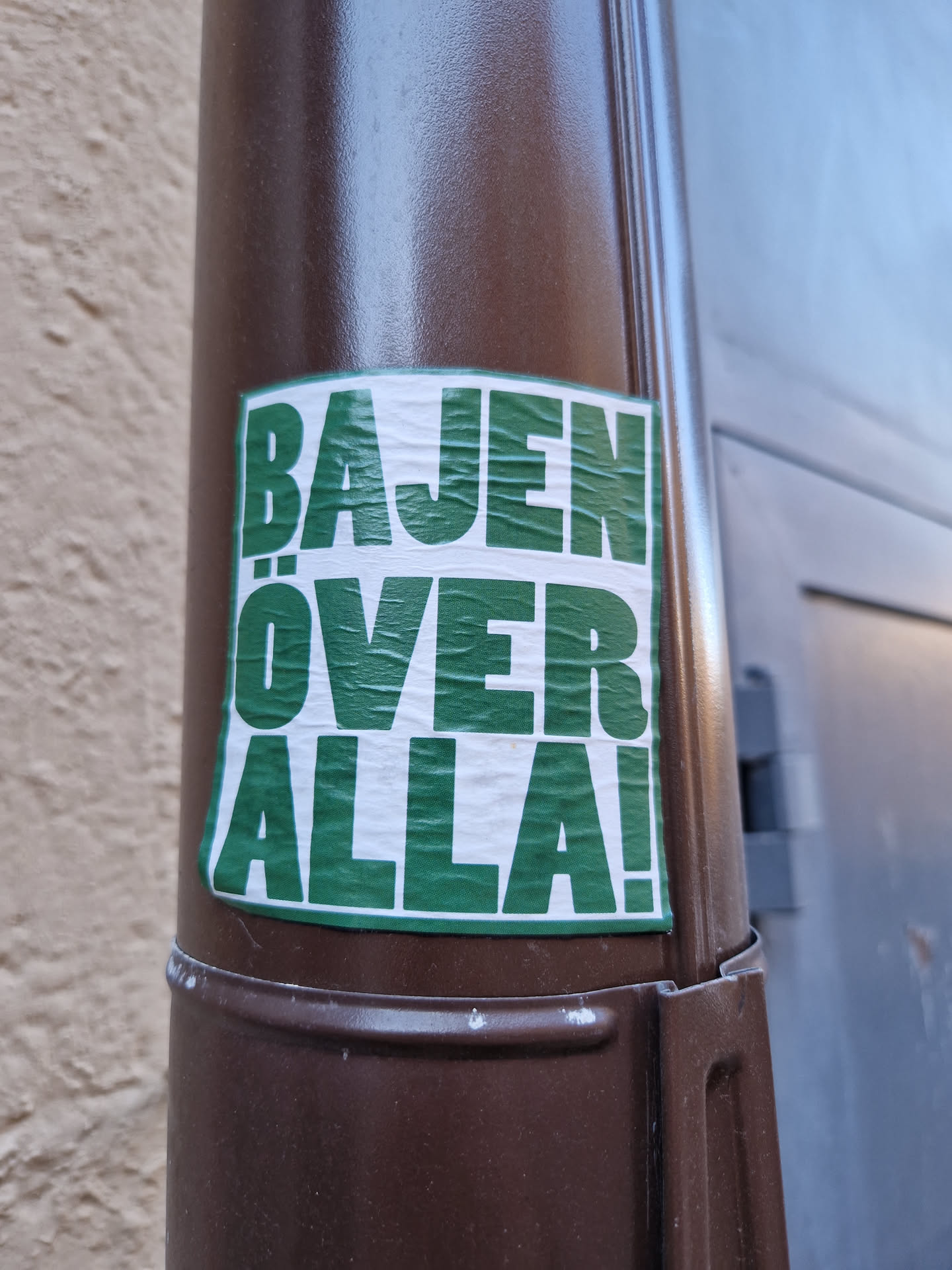 A green sticker with white text on a brown pole. The text says "BAJEN ÖVER ALLA!"