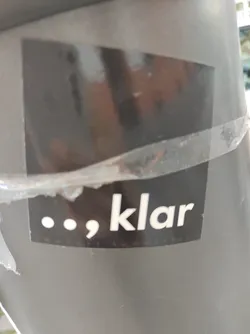 A dark gray square sticker with the word 'klar' in white text and two dots and a comma before it.  The sticker is attached to a gray surface.  The text appears to be a brand name or logo.  The overall aesthetic is clean and modern.