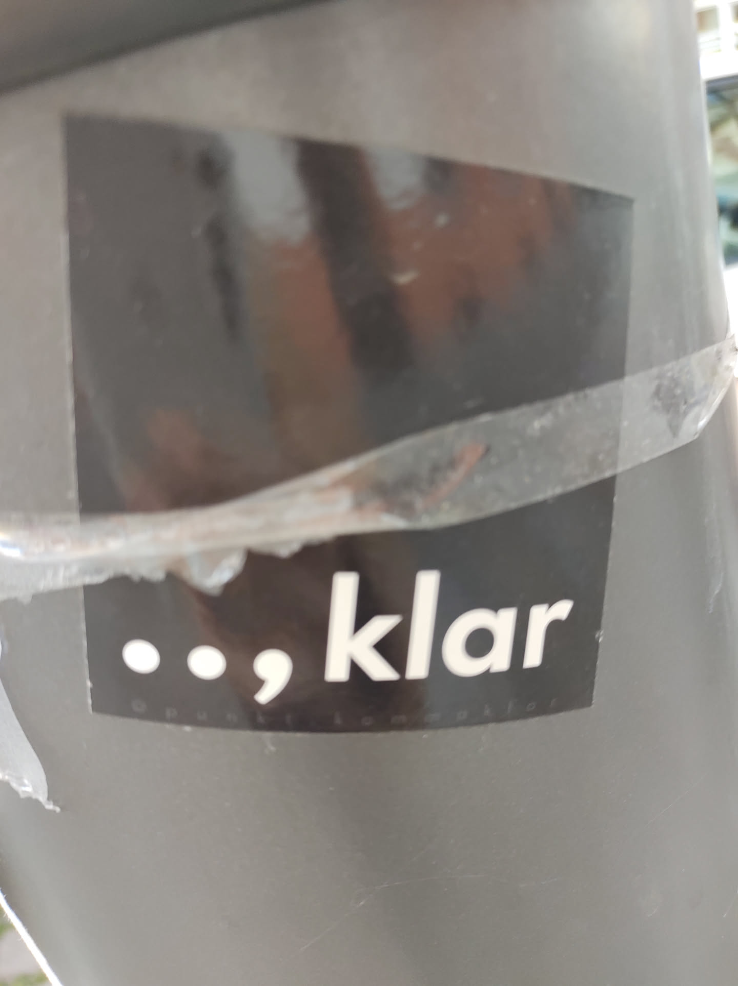 A dark gray square sticker with the word 'klar' in white text and two dots and a comma before it.  The sticker is attached to a gray surface.  The text appears to be a brand name or logo.  The overall aesthetic is clean and modern.