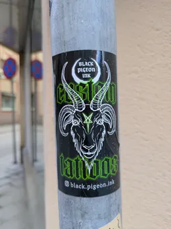 A sticker for Black Pigeon Ink, a tattoo shop, is affixed to a gray pole. The sticker is predominantly black with green and white details. The main image depicts a ram-like creature with a pentagram on its forehead. The text on the sticker reads "Black Pigeon Ink", "Custom Tattoos" and their Instagram handle.