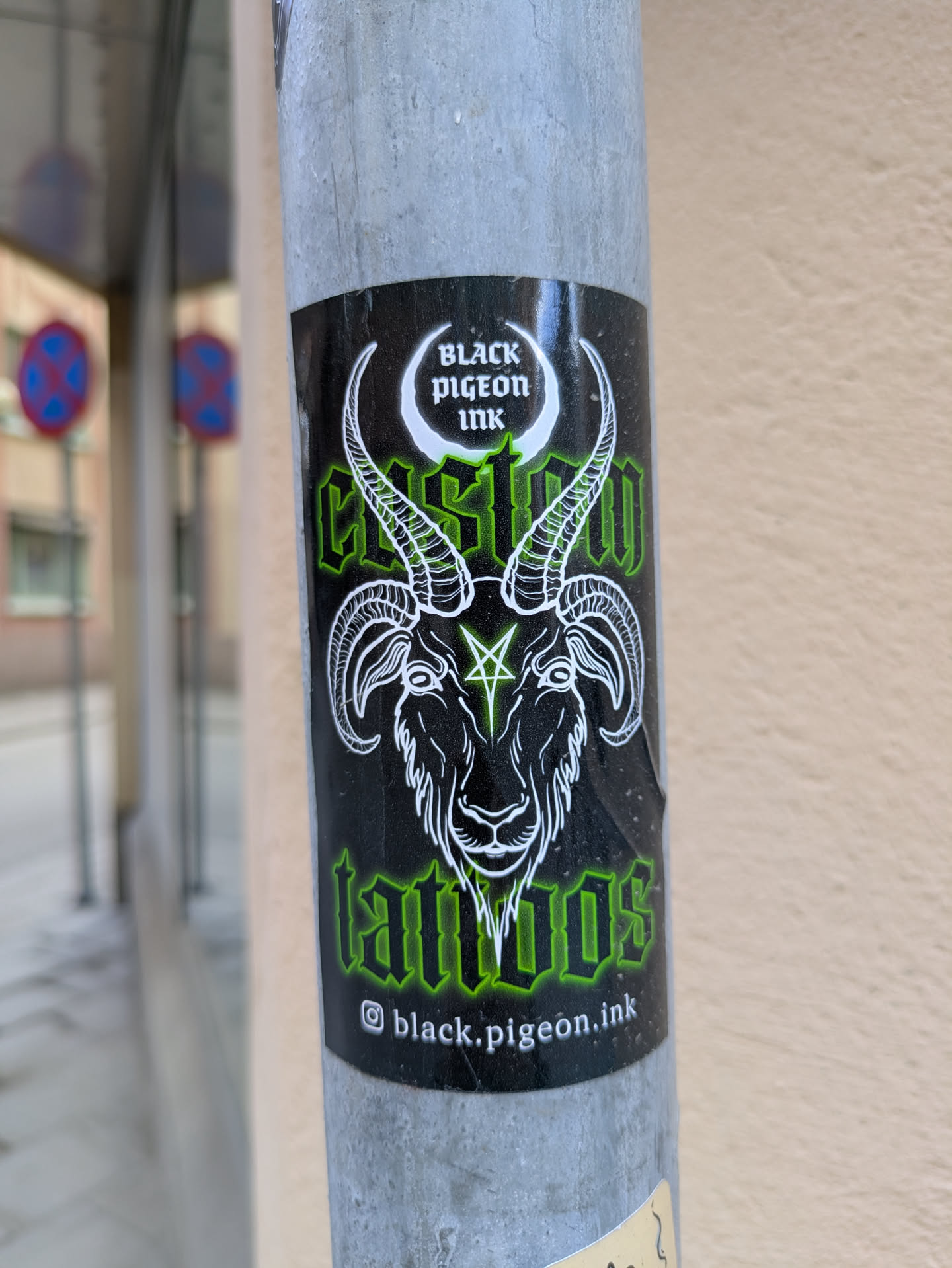 A sticker for Black Pigeon Ink, a tattoo shop, is affixed to a gray pole. The sticker is predominantly black with green and white details. The main image depicts a ram-like creature with a pentagram on its forehead. The text on the sticker reads "Black Pigeon Ink", "Custom Tattoos" and their Instagram handle.