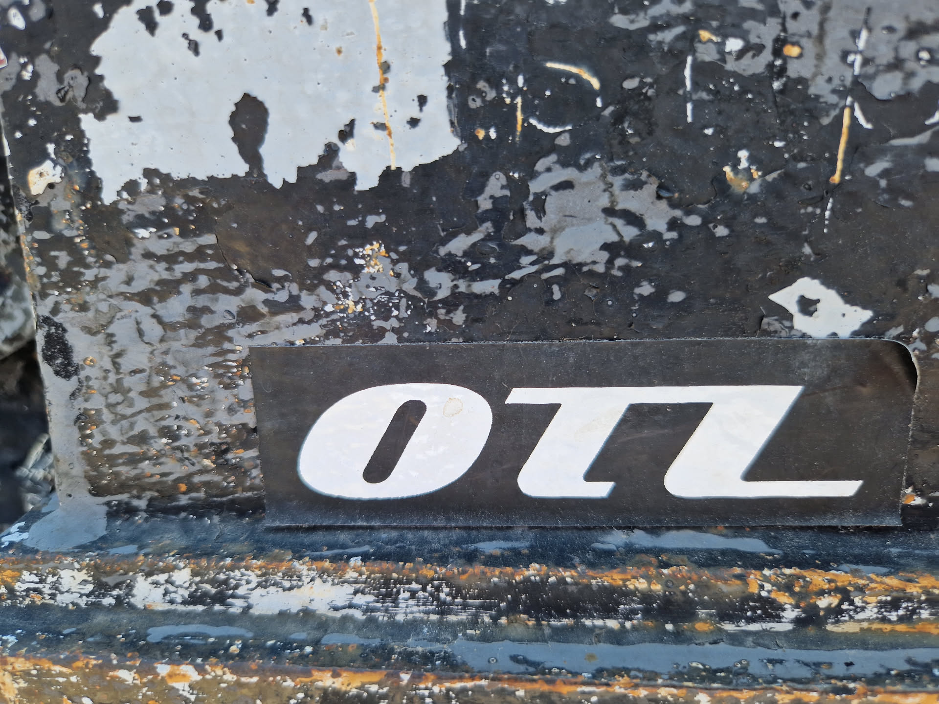 A close-up shot of a black sticker with the letters "OTL" in white. The sticker is affixed to a heavily textured, weathered surface, possibly metal, with peeling paint and various markings.
