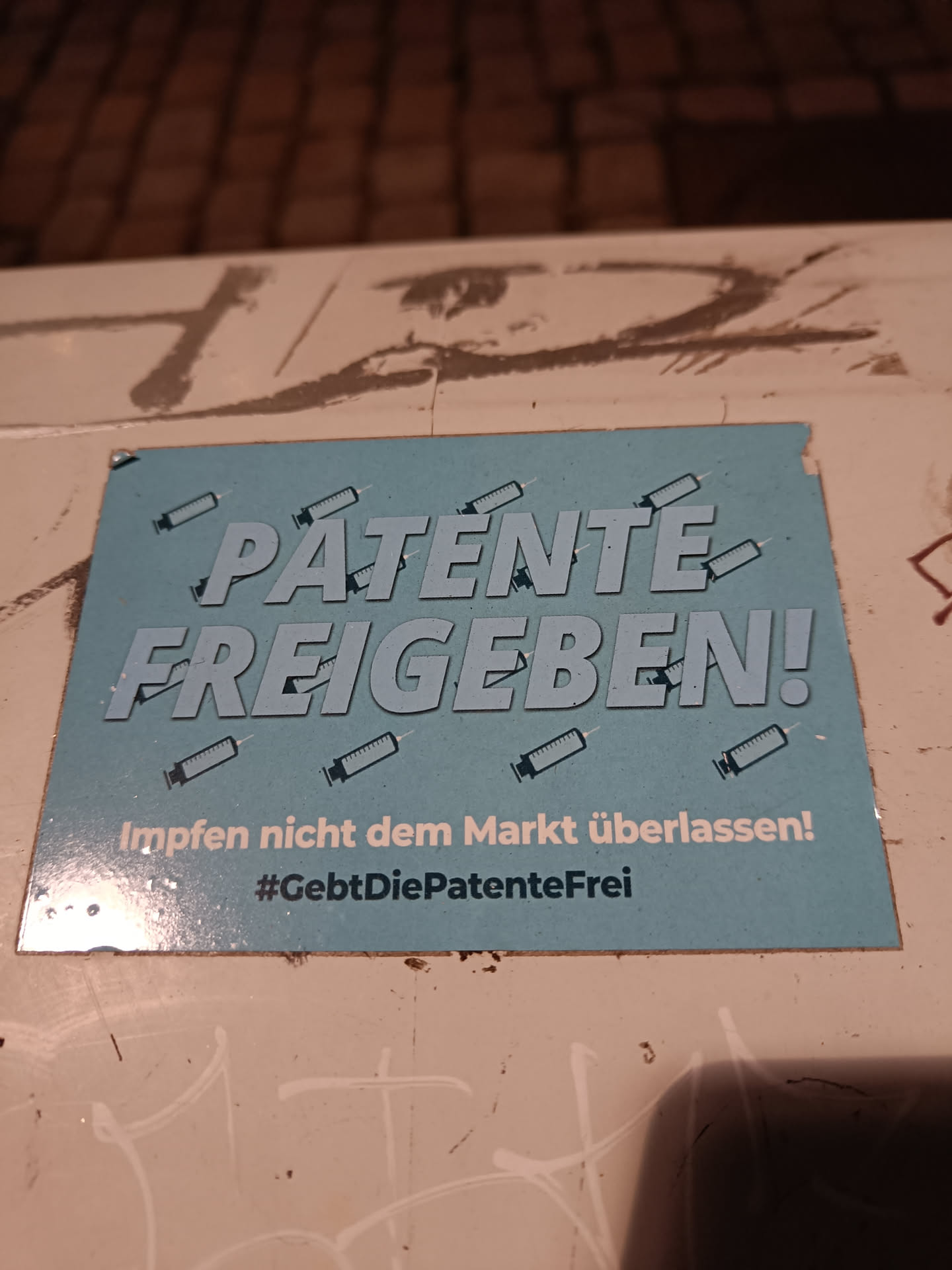 A sticker advocating for the release of vaccine patents. The sticker is light blue with the text "PATENTE FREIGEBEN!" in bold, capital letters.  Below it, in smaller text, it says "Impfen nicht dem Markt überlassen!" (#GebtDiePatenteFrei).  Several images of syringes are also on the sticker.