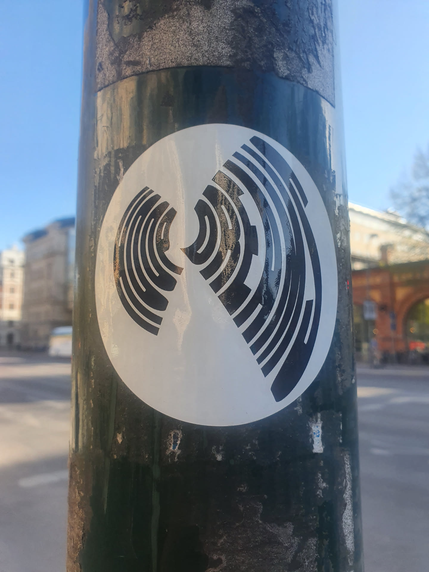 A round sticker with an abstract, geometric design in black and white.  The design resembles two mirrored swirling patterns, creating a sense of movement and depth. The sticker is affixed to a weathered green pole, suggesting it has been there for some time.