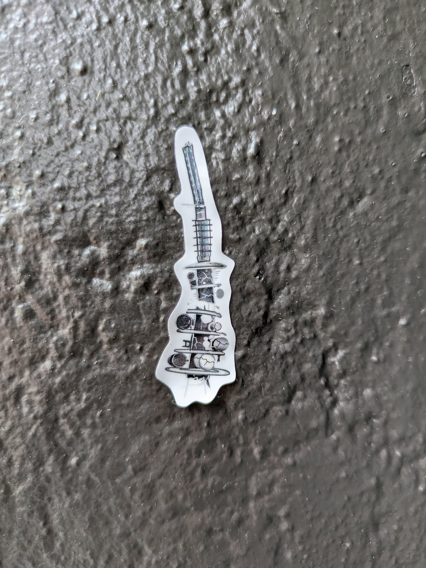 A vertically elongated sticker featuring a detailed black and white line drawing. The artwork depicts a complex, almost surreal structure that resembles a cross-section of a futuristic machine or a deconstructed architectural element. It includes a tower-like top section with linear details, possibly representing a spring or antenna, leading down to multiple platforms. These platforms hold various spherical or orb-like objects, some appearing cracked or broken, suggesting decay or fragmentation. The overall aesthetic is one of intricate, almost blueprint-like design with a distressed or post-apocalyptic feel. The sticker is placed on a rough, dark grey or black textured surface.
