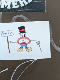 A cartoonish character sticker on a wall. It's a mouth with eyes and a black top hat with a red stripe, wearing blue trousers and brown shoes, holding a sign that says "BocMan" in one hand and a cane in the other.