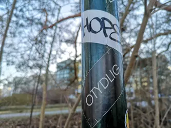 The central sticker is a black, rectangular shape, oriented diagonally on a dark green pole. It features the word "OTYDLIG" in clear, sans-serif white capital letters, running from the bottom left to the top right. The sticker has a subtle white outline or border, and its surface shows minor scuffs and dust, indicating it's an outdoor application. The background of the sticker is a solid, matte black.