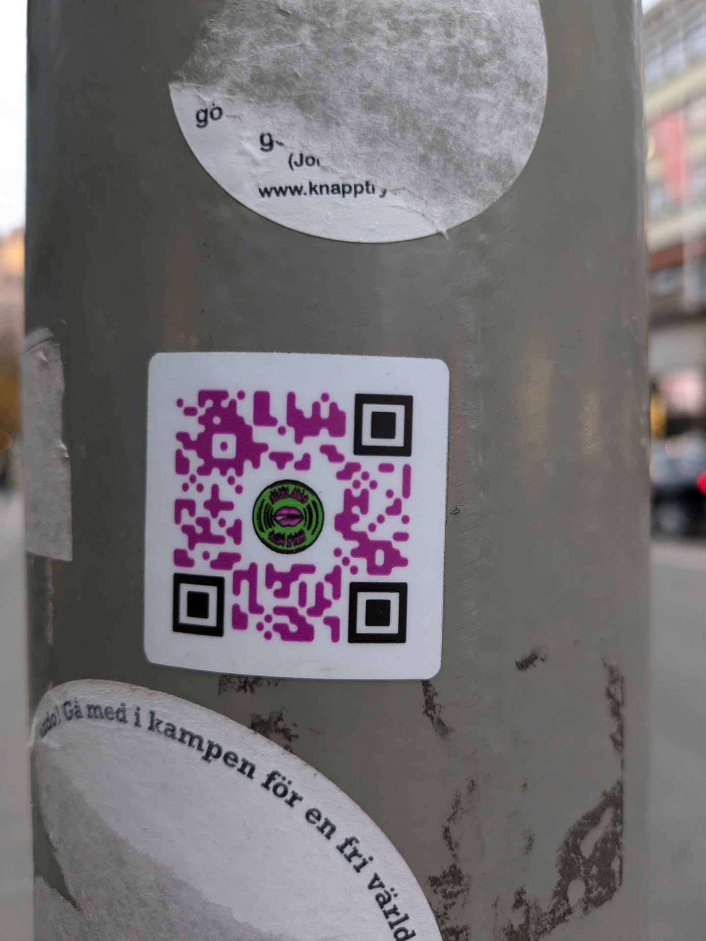 A sticker with a QR code in the center.  The QR code has a small logo in the middle and purple accents. The text around the bottom of the sticker says "Gå med i kampen för en fri värld" which translates from Swedish to "Join the fight for a free world".  There's another sticker above it with partial text visible: "www.knappt"