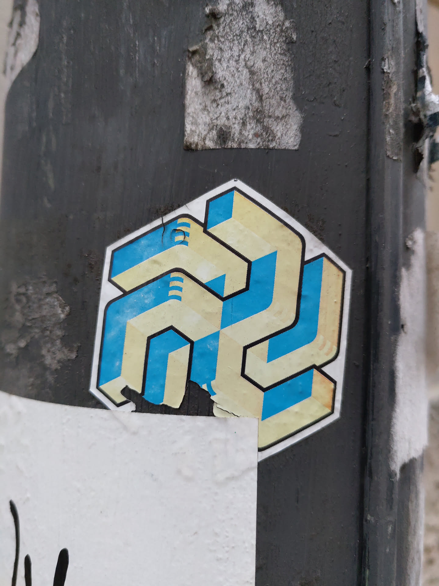 Close-up view of a sticker featuring an abstract design. The design is primarily composed of geometric shapes in beige and teal, creating a three-dimensional effect. The sticker is affixed to a dark grey pole, and a portion of the sticker is slightly damaged or peeled.