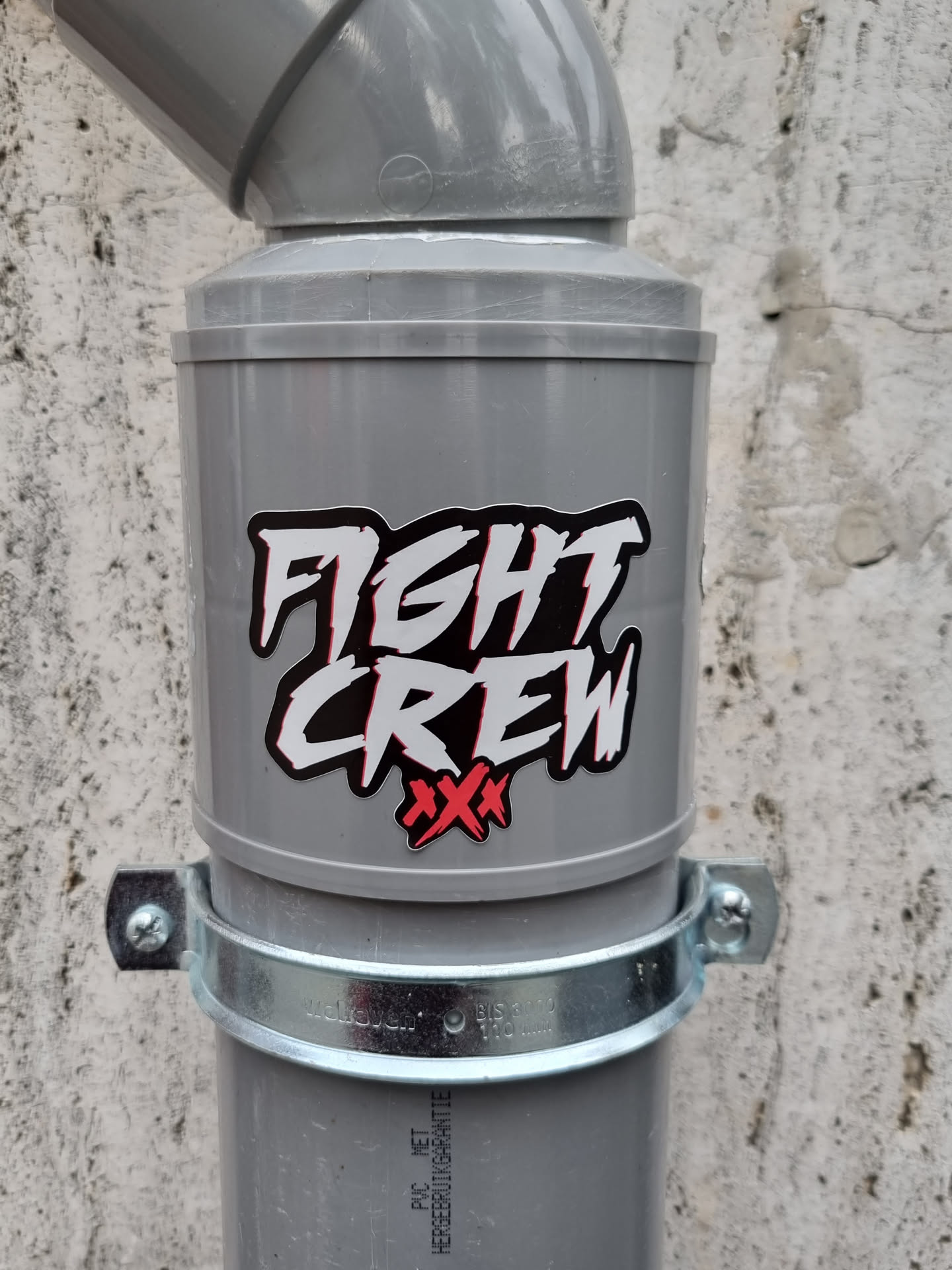 A sticker with the text "FIGHT CREW" in stylized lettering and two red crosses is affixed to a grey pipe. The sticker appears to be vinyl and is placed near the top of the pipe.