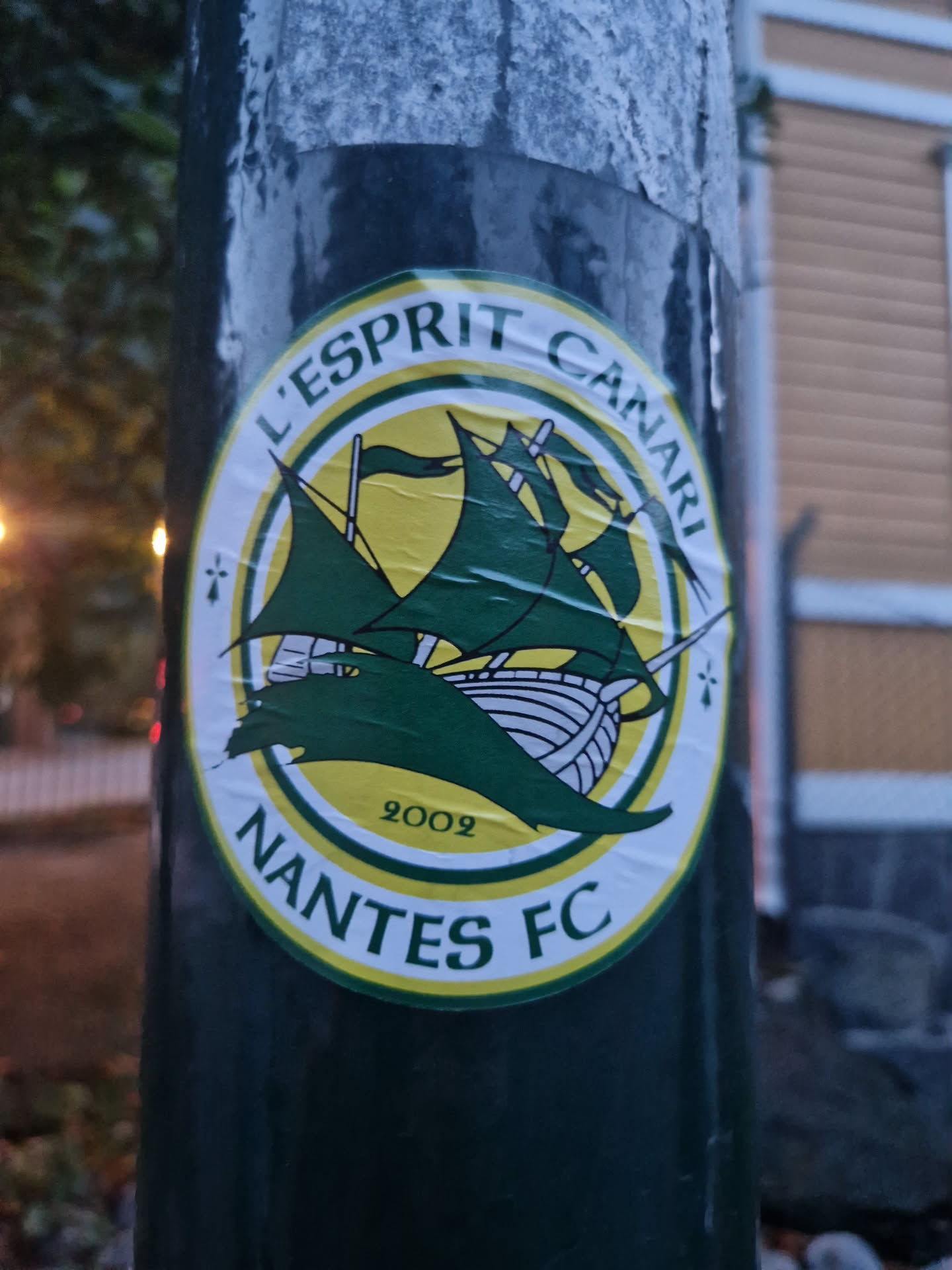 A green and yellow circular sticker featuring the Nantes FC logo. The logo includes a ship, the text "L'Esprit Canari" and "Nantes FC", and the year 2002. The sticker is affixed to a dark green pole.