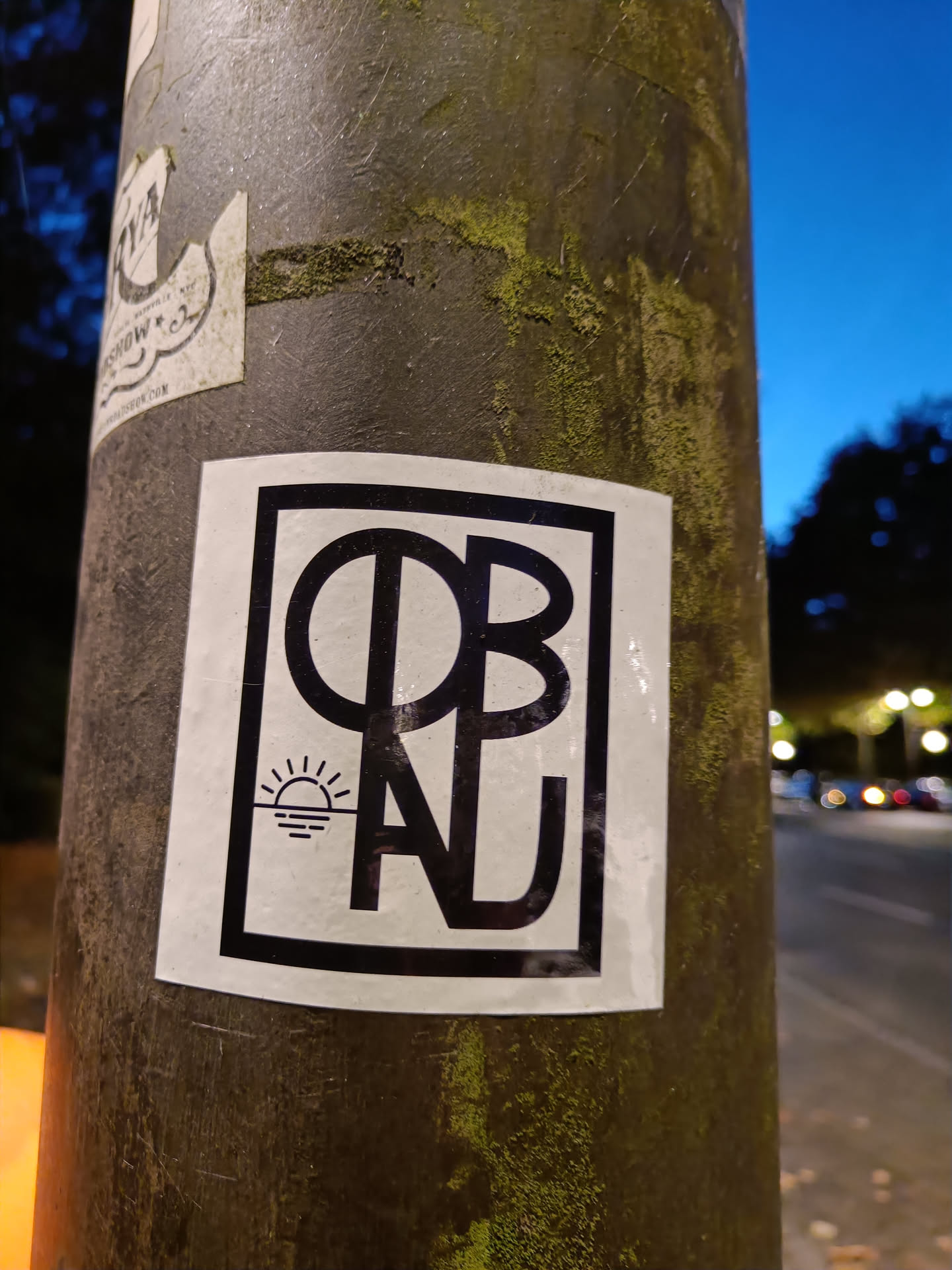 A black and white sticker featuring a stylized rectangular frame around the bold, uppercase initials "OBAL".  A small image of a setting sun over water is present within the frame, above the letters. The sticker is affixed to a weathered grey metal pole outdoors.
