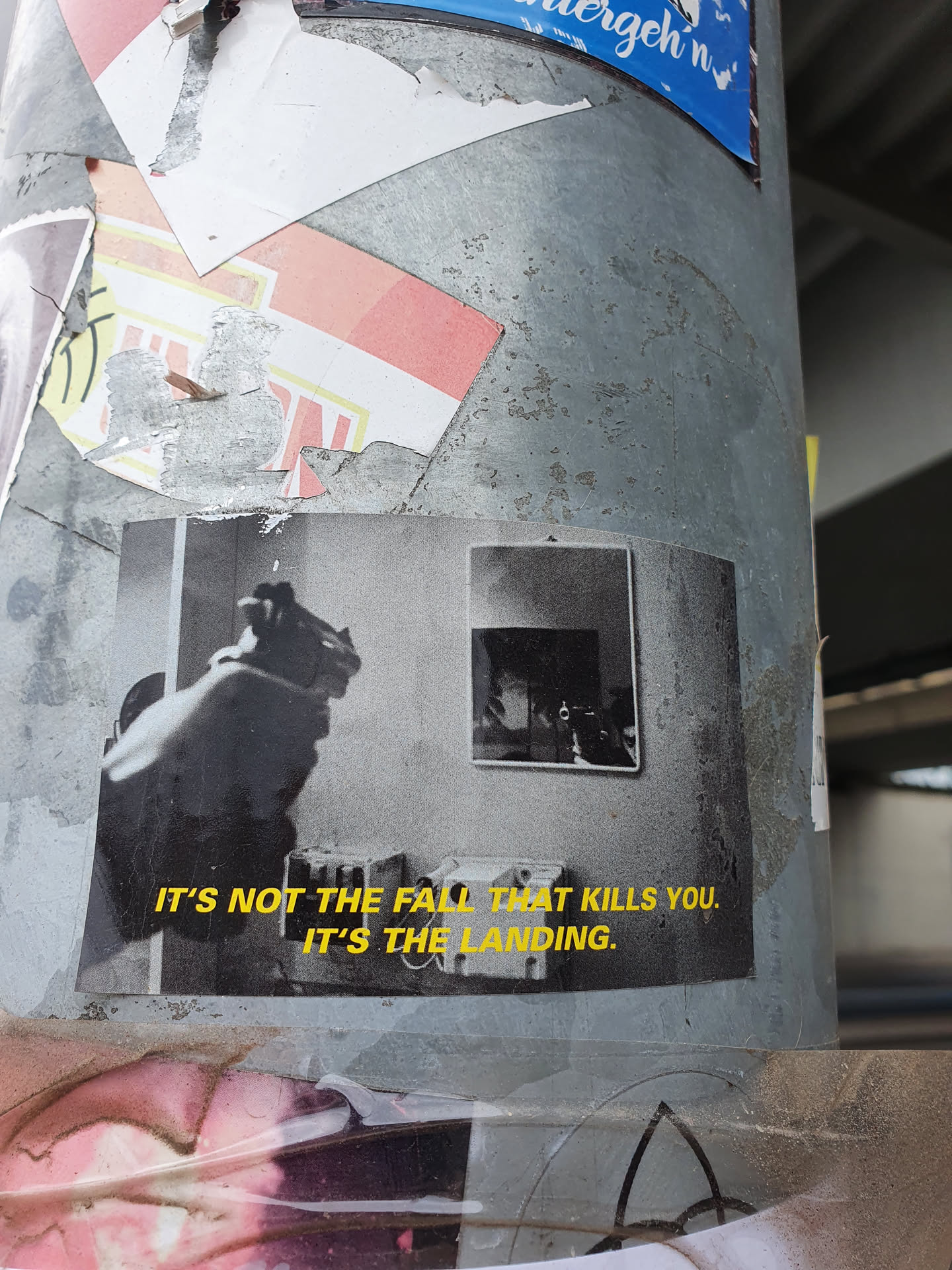 A black and white photo sticker depicting a hand holding a gun, with a smaller framed photo in the background.  The text "It's not the fall that kills you. It's the landing." is printed below the image in yellow.
