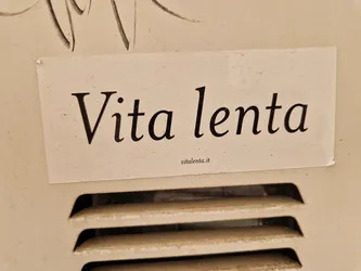A beige sticker with the text "Vita lenta" (slow life in Italian) on it. The sticker is attached to a beige wall.  There is a small website address visible at the bottom of the sticker.